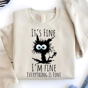 May include: A beige sweatshirt with a black cat illustration and the text "It's fine, I'm fine, everything is fine".