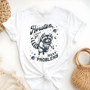 Op de afbeelding: Wit T-shirt met een wasbeer-astronaut ontwerp en de tekst "Houston, I have 50 many problems." De afbeelding bevat planeten en sterren. Het shirt is in de taille geknoopt.