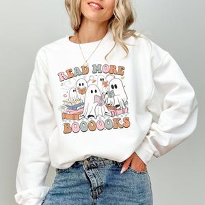 Può includere: Felpa bianca con una grafica colorata che recita "Read More Booooooks". Il design presenta fantasmi dei cartoni animati che leggono libri. Il testo e le illustrazioni sono in tonalità arancioni, rosa, verdi e blu.