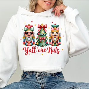 Op de afbeelding: Witte hoodie met een kleurrijke afbeelding van twee notenkrakerfiguren en een kerstboom. De tekst "Y'all are Nuts" is in rode letters gedrukt. Het ontwerp bevat rode en gouden accenten, perfect voor de feestdagen.