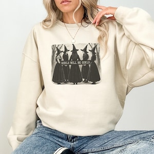 Può includere: Felpa color crema con grafica in bianco e nero di cinque streghe e la scritta "GIRLS WILL BE GIRLS." Le streghe indossano abiti lunghi e cappelli a punta. La felpa è in stile casual.