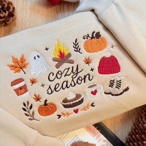 Puede incluir: Una sudadera beige con bordados de motivos oto&ntilde;ales que incluyen una fogata, un fantasma, calabazas, hojas de arce, un gorro rojo, una taza de caf&eacute;, un pastel y botas, con el texto "cozy season" bordado en letras cursivas de color marr&oacute;n.