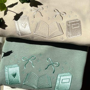 Op de afbeelding: Twee sweatshirts, één beige en één groen, geborduurd met boek- en strikdesigns. Het beige sweatshirt heeft een hart op één boek. Het groene sweatshirt heeft een hart op één boek. Beide hebben open boeken en steraccenten.