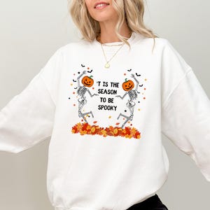 Op de afbeelding: Witte sweatshirt met een Halloween-thema. Het ontwerp toont twee dansende skeletten met pompoenhoofden, omringd door herfstbladeren en de tekst "'T IS THE SEASON TO BE SPOOKY".