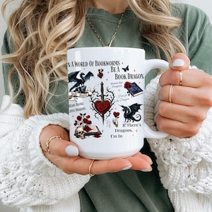 Op de afbeelding: Witte keramische mok met de tekst "IN A WORLD OF BOOKWORMS + BE A BOOK DRAGON". De mok heeft illustraties van draken, boeken, rozen en een zwaard. De mok wordt door een persoon vastgehouden.