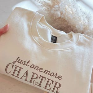 Peut inclure: Un sweat-shirt à col rond de couleur crème avec le texte brodé "just one more CHAPTER" en fil marron. Les mots "just one more" sont en écriture cursive, tandis que "CHAPTER" est en police serif grasse avec un petit cœur.