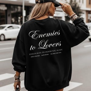 Op de afbeelding: Zwart sweatshirt met de tekst "Enemies to Lovers" in een sierlijk lettertype. Daaronder staan de zinnen "EVEN IN RUIN, I'LL CHOOSE YOU AGAIN", "ONLY ONE BED", "SLOW BURN" en "I'LL KILL FOR YOU".