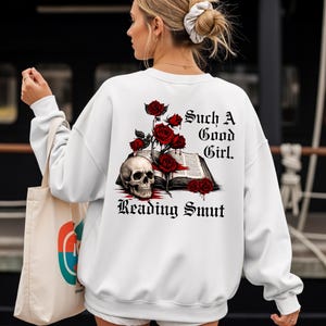 Op de afbeelding: Witte sweatshirt met een afbeelding op de achterkant met een schedel, rode rozen en een open boek. De tekst luidt: "Such A Good Girl. Reading Smut."