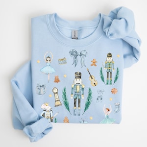 Op de afbeelding: Een lichtblauwe sweatshirt met een aquarelontwerp van het ballet De Notenkraker. Het ontwerp bevat een ballerina, notenkrakers, een muizenkoning en andere feestelijke elementen. De sweatshirt is gemaakt van zacht materiaal.