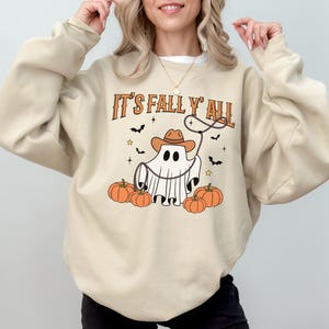 Może przedstawiać: Beżowa bluza z napisem "IT'S FALL Y'ALL" w kolorze pomarańczowym. Grafika przedstawia ducha w kapeluszu kowbojskim i z lassem, otoczonego dyniami, nietoperzami i gwiazdami. Zabawny, sezonowy design.