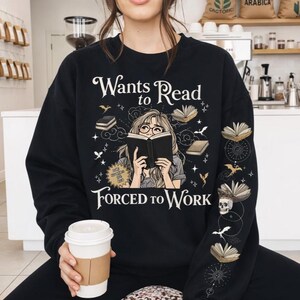 Könnte beinhalten: Schwarzes Sweatshirt mit dem Text "Wants to Read, Forced to Work" und einer Grafik einer Person, die ein Buch liest. Das Design umfasst Illustrationen von Büchern, Sternen und einem Totenkopf. Die Person hält eine weiße Kaffeetasse.