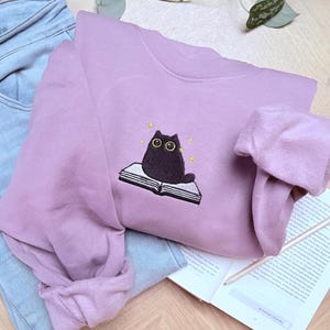 Op de afbeelding: Mauvekleurige sweatshirt met een borduursel van een zwarte kat die op een open boek zit. De kat heeft gele ogen en is omringd door kleine gele sterren. De sweatshirt ligt op een lichtblauwe jeans.
