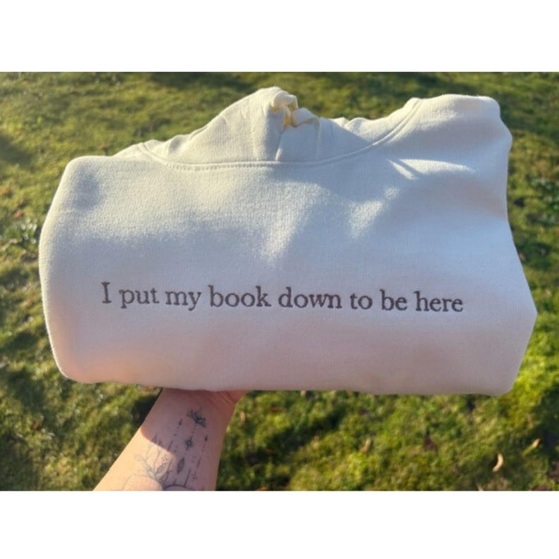 Op de afbeelding: Een beige sweatshirt met de tekst "I put my book down to be here" in het zwart. De sweatshirt is opgevouwen, met een deel van een spijkerbroek zichtbaar. Het Gildan-label is zichtbaar.