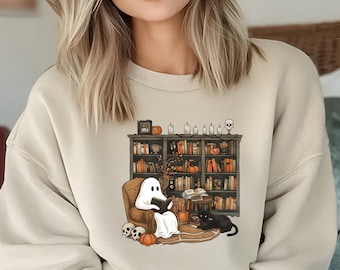 Könnte beinhalten: Beiges Sweatshirt mit einem Halloween-Motiv. Das Design zeigt einen Geist, der in einem Sessel vor einem Bücherregal mit Büchern, Kürbissen und Kerzen liest. Eine schwarze Katze sitzt daneben. Schädel und Kürbisse sind ebenfalls vorhanden.