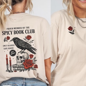 Peut inclure: Un t-shirt beige avec un corbeau noir perché sur un crâne, entouré de roses rouges et de livres. Le texte "Proud Member of the Spicy Book Club" est imprimé sur le t-shirt, ainsi que les mots "Smut Readers' Safe Haven" et "Meet Weekly".