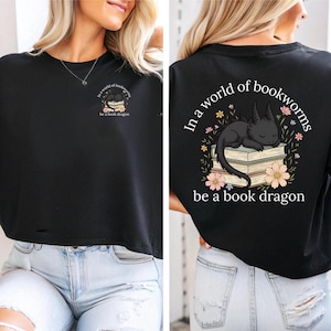 Pode incluir: Camiseta preta cropped com um gráfico de um dragão preto dormindo em cima de uma pilha de livros. O texto "In a world of bookworms be a book dragon" está impresso na parte de trás da camiseta.