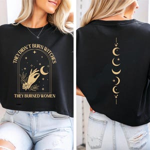Può includere: Top corto nero con testo e grafica dorati. Il davanti presenta la scritta "THEY DIDN'T BURN WITCHES THEY BURNED WOMEN" all'interno di un disegno ad arco con una mano, luna, stelle ed elementi floreali. Il retro mostra una linea verticale di lune crescenti e stelle.