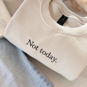 Op de afbeelding: Crèmekleurige sweatshirt met de zwarte tekst "Not today". De sweatshirt is opgevouwen en naast een lichtblauwe jeans geplaatst. Het Gildan-merkteken is zichtbaar.