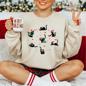 Może przedstawiać: Beżowa bluza z świątecznym wzorem czarnych kotów w czapkach Świętego Mikołaja, otoczonych płatkami śniegu i ozdobami. Bluza ma okrągły dekolt i długie rękawy. W dłoni trzymany jest biały kubek z napisem "MERRY CHRISTMAS".