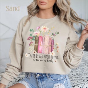 Peut inclure: Un sweat-shirt beige avec un motif floral et une pile de livres. Le texte "THERE IS NO SUCH THING as too many books" est imprimé sur le devant du sweat-shirt.