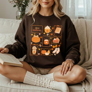 Può includere: Una felpa marrone con una grafica a tema autunnale. La grafica include una zucca, una tazza di caffè, una candela, una torta e il testo "Hello Autumn". Il design è nei toni dell'arancione, marrone e bianco.