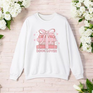 Peut inclure: Sweat-shirt blanc avec un motif rose d'une pile de livres liés par un ruban rose et le texte "BOOK LOVER".