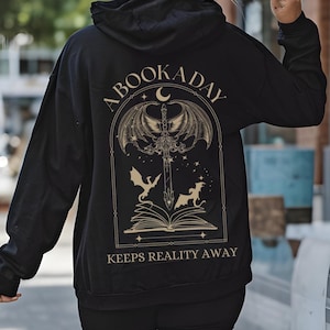 Könnte beinhalten: Schwarzer Kapuzenpullover mit einer goldenen Grafik eines Buches, eines Schwertes und Drachen. Der Text "A Book A Day Keeps Reality Away" ist auf der Rückseite aufgedruckt.