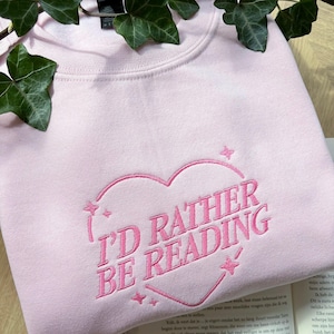 Op de afbeelding: Lichtroze sweatshirt met de geborduurde tekst "I'D RATHER BE READING" in een hartvorm. Het ontwerp is in bijpassend roze garen. Het sweatshirt ligt op een boek.