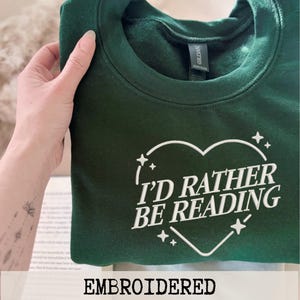 Può includere: Una felpa verde scuro con la scritta "I'D RATHER BE READING" all'interno di un cuore. La parola "EMBROIDERED" è in basso nell'immagine. La felpa è in stile casual.