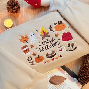 Geborduurde beige sweatshirt met het opschrift 'Cozy Season' en een herfstig borduurontwerp van een kampvuur, pompoenen, spookje, pumpkin spice koffie, warme chocolademelk, herfstbladeren, taart, gebreide muts en winterlaarsjes. De sweater ligt op een open boek met dennenappels, kaarslicht en herfstdecoratie. Perfecte herfsttrui voor Halloween, cozy season en herfstliefhebbers.
