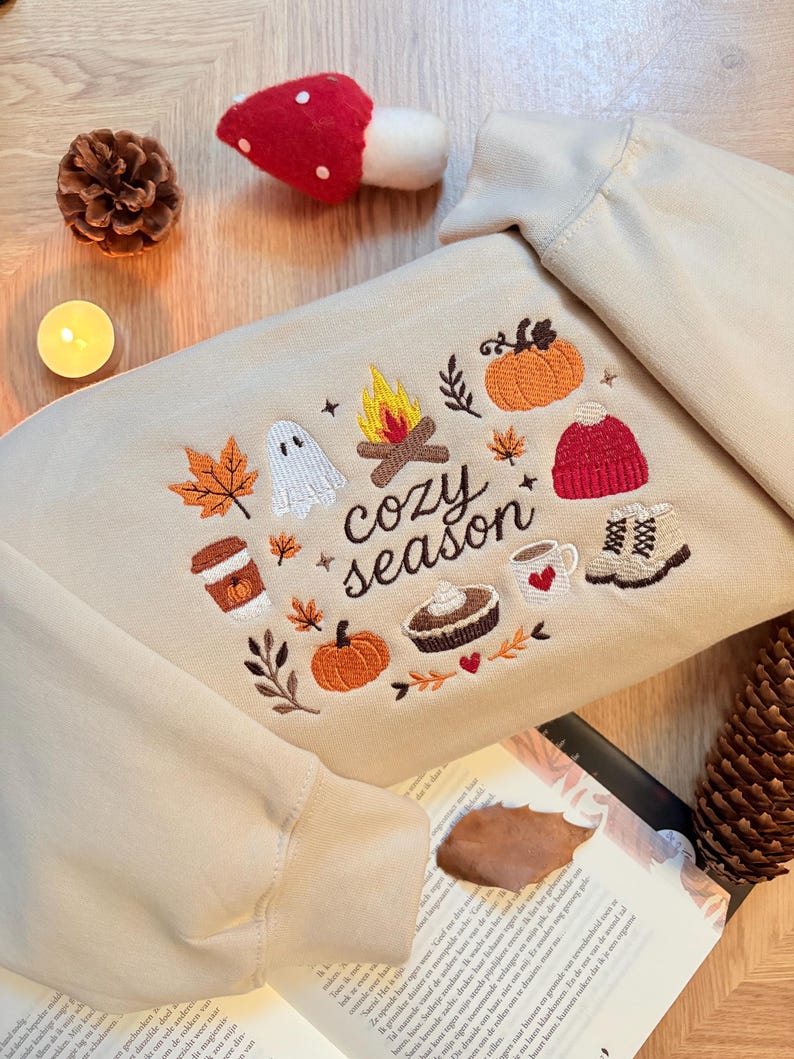 Geborduurde beige sweatshirt met het opschrift 'Cozy Season' en een herfstig borduurontwerp van een kampvuur, pompoenen, spookje, pumpkin spice koffie, warme chocolademelk, herfstbladeren, taart, gebreide muts en winterlaarsjes. De sweater ligt op een open boek met dennenappels, kaarslicht en herfstdecoratie. Perfecte herfsttrui voor Halloween, cozy season en herfstliefhebbers.