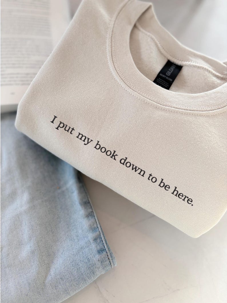 May include: A beige sweatshirt with the text "I put my book down to be here." The sweatshirt is folded and placed on top of a pair of light blue jeans. The sweatshirt has a crew neck and long sleeves.