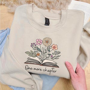 Op de afbeelding: Beige sweatshirt met een bloemenontwerp boven een open boek. Het ontwerp bevat roze, perzik en gele bloemen met groene bladeren. De tekst "One more chapter" staat onder het boek. De sweatshirt is op een blauwe jeans gevouwen.
