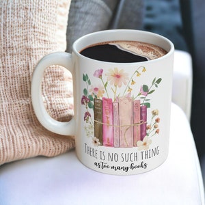 Può includere: Tazza in ceramica bianca con caffè nero al suo interno. La tazza ha un design floreale con libri rosa e marroni e il testo "THERE IS NO SUCH THING as too many books".