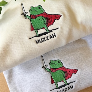 Op de afbeelding: Twee sweatshirts, één crème en één lichtgrijs, elk voorzien van een geborduurde afbeelding van een groene kikker met een rode cape die een zilveren zwaard vasthoudt. Onder de kikker is het woord HUZZAH in zwarte draad op beide kledingstukken geborduurd.