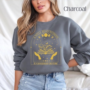 Puede incluir: Una sudadera gris antracita con un diseño gráfico dorado. El gráfico presenta un libro con flores, manos y el texto "A well read woman is a dangerous creature".