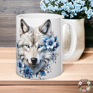 Op de afbeelding: Witte keramische mok met een afbeelding van een witte wolf met blauwe ogen, omringd door blauwe bloemen.