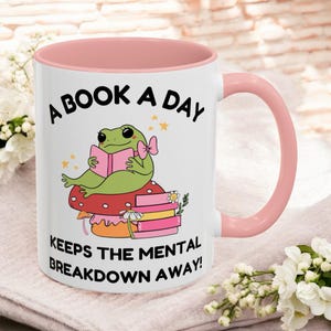 Può includere: Una tazza in ceramica bianca con un bordo e un manico rosa. La tazza presenta una rana verde seduta su un fungo con una pila di libri. Il testo sulla tazza recita "A book a day keeps the mental breakdown away!"