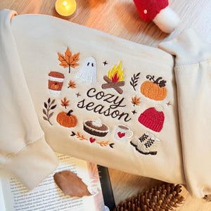Geborduurde beige sweatshirt met het opschrift 'Cozy Season' en een herfstig borduurontwerp van een kampvuur, pompoenen, spookje, pumpkin spice koffie, warme chocolademelk, herfstbladeren, taart, gebreide muts en winterlaarsjes. De sweater ligt op een open boek met dennenappels, kaarslicht en herfstdecoratie. Perfecte herfsttrui voor Halloween, cozy season en herfstliefhebbers.