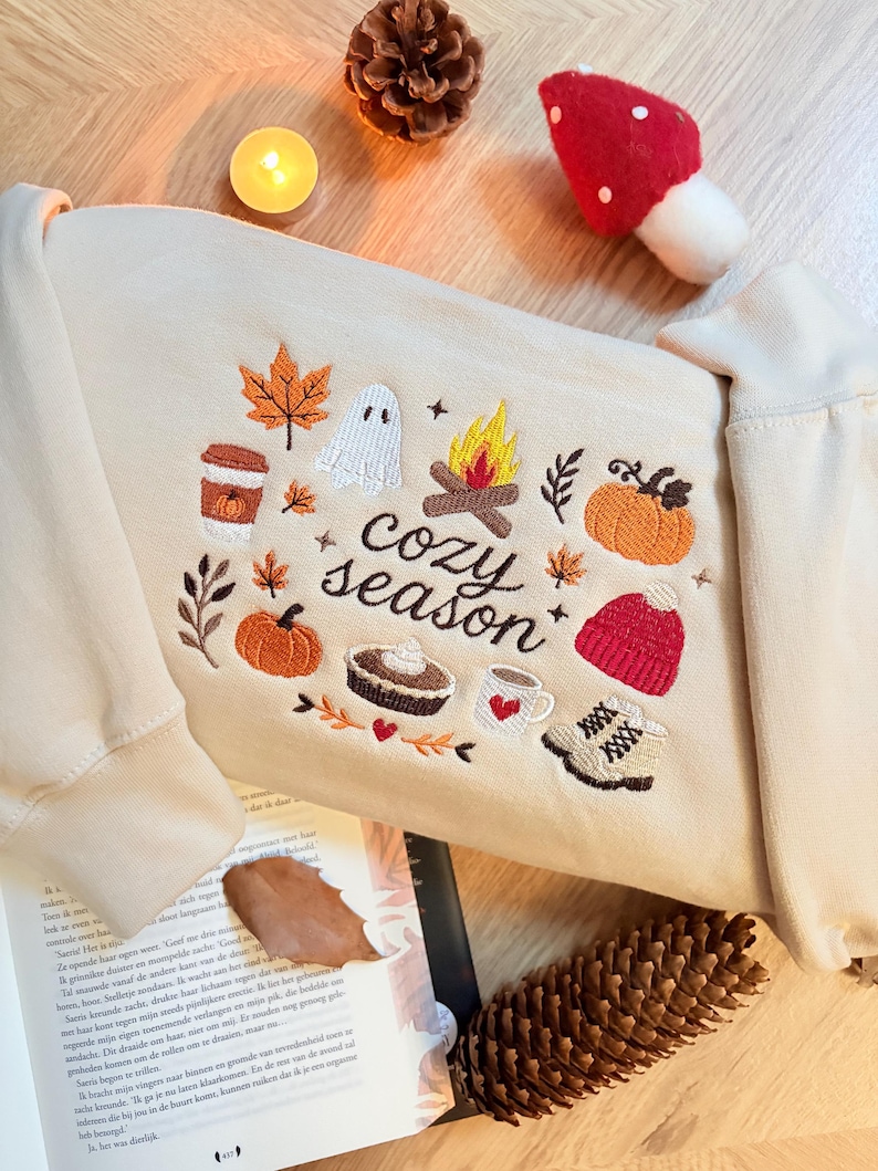 Geborduurde beige sweatshirt met het opschrift 'Cozy Season' en een herfstig borduurontwerp van een kampvuur, pompoenen, spookje, pumpkin spice koffie, warme chocolademelk, herfstbladeren, taart, gebreide muts en winterlaarsjes. De sweater ligt op een open boek met dennenappels, kaarslicht en herfstdecoratie. Perfecte herfsttrui voor Halloween, cozy season en herfstliefhebbers.