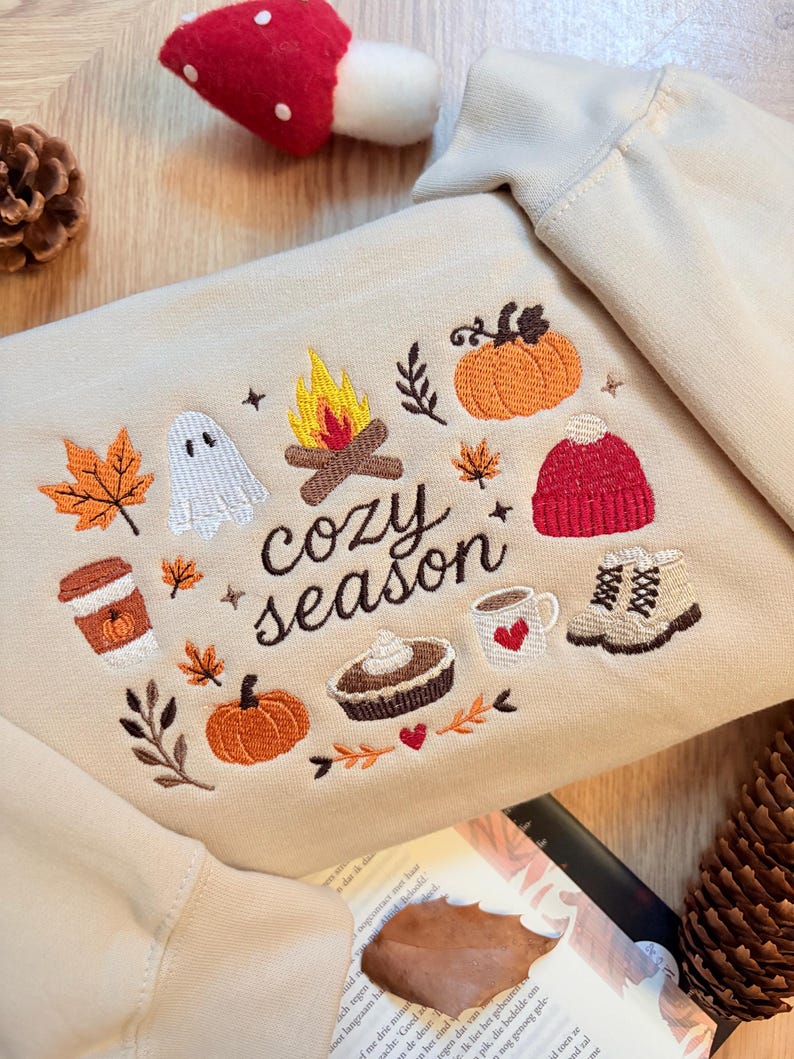 Puede incluir: Una sudadera beige con bordados de motivos oto&ntilde;ales que incluyen una fogata, un fantasma, calabazas, hojas de arce, un gorro rojo, una taza de caf&eacute;, un pastel y botas, con el texto "cozy season" bordado en letras cursivas de color marr&oacute;n.
