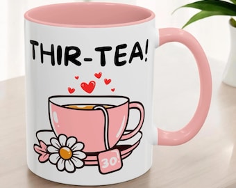 Op de afbeelding: Witte keramische mok met een roze rand en handvat. De mok heeft een roze theekopje en schoteltje met een theezakje waarop "30!" staat en de tekst "THIR-TEA!" in zwart.