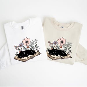 Op de afbeelding: Twee sweatshirts, één wit en één beige, elk met een afbeelding van een zwarte kat die op een open boek slaapt, omringd door roze, paarse en groene bloemen. Het ontwerp is gecentreerd op de borst.