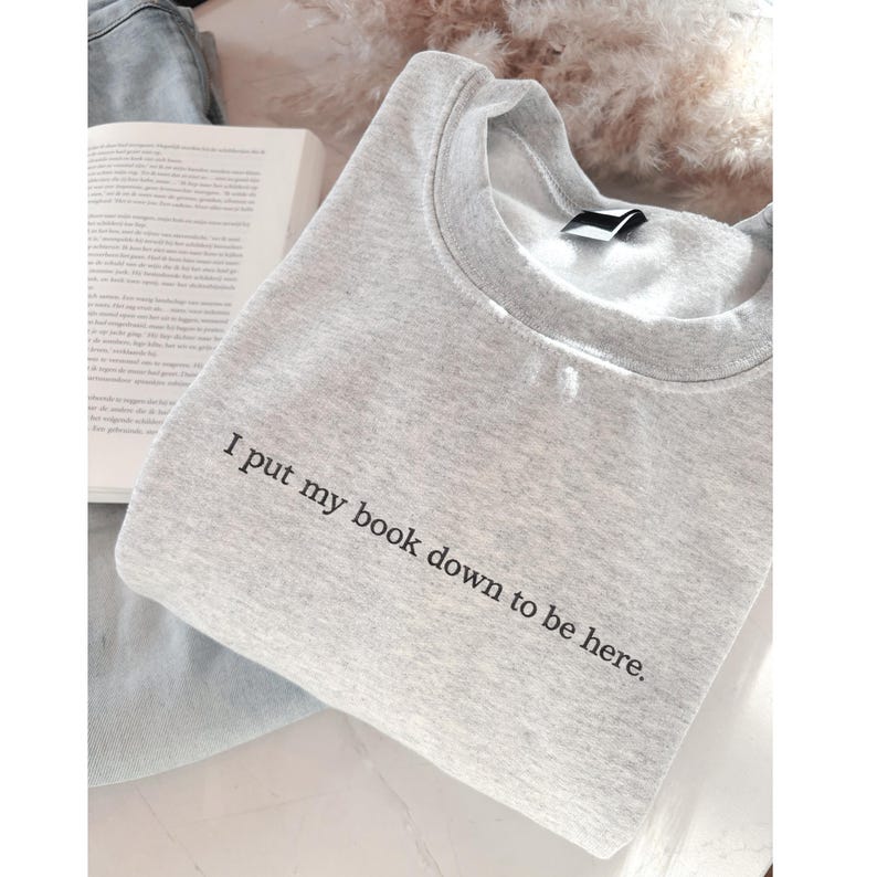 May include: A light gray sweatshirt with the text "I put my book down to be here." The crew neck sweatshirt is folded and placed next to an open book. The background is a soft, neutral color.