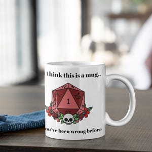 May include: White ceramic mug with a handle. The mug features a red, geometric design with the number 1, a skull, and floral accents. Text on the mug reads "I think this is a mug.. You've been wrong before."