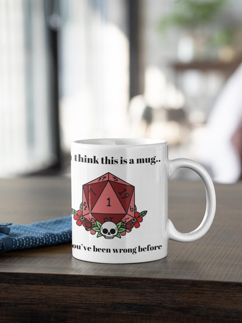 May include: White ceramic mug with a handle. The mug features a red, geometric design with the number 1, a skull, and floral accents. Text on the mug reads "I think this is a mug.. You've been wrong before."