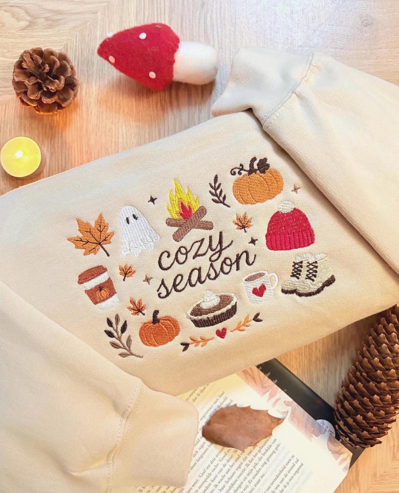 Geborduurde beige sweatshirt met het opschrift 'Cozy Season' en een herfstig borduurontwerp van een kampvuur, pompoenen, spookje, pumpkin spice koffie, warme chocolademelk, herfstbladeren, taart, gebreide muts en winterlaarsjes. De sweater ligt op een open boek met dennenappels, kaarslicht en herfstdecoratie. Perfecte herfsttrui voor Halloween, cozy season en herfstliefhebbers.