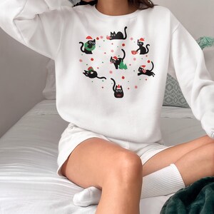 K&ouml;nnte beinhalten: Wei&szlig;er Pullover mit einem festlichen Design mit schwarzen Katzen, die Weihnachtsm&uuml;tzen tragen und weihnachtliche Gegenst&auml;nde wie einen Kranz, einen Baum und ein Geschenk halten. Die Katzen sind von Schneeflocken und roten Sternen umgeben.