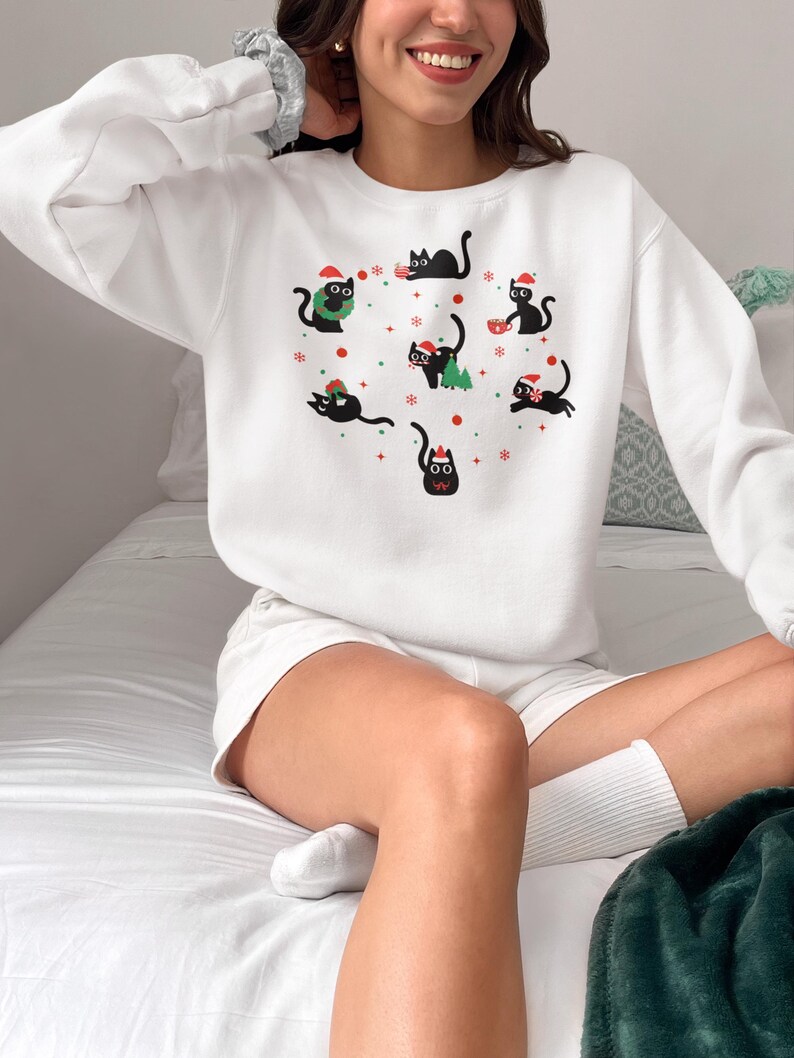 K&ouml;nnte beinhalten: Wei&szlig;er Pullover mit einem festlichen Design mit schwarzen Katzen, die Weihnachtsm&uuml;tzen tragen und weihnachtliche Gegenst&auml;nde wie einen Kranz, einen Baum und ein Geschenk halten. Die Katzen sind von Schneeflocken und roten Sternen umgeben.