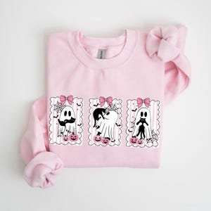 Op de afbeelding: Een lichtroze sweatshirt met drie ingelijste spookillustraties. Elke geest heeft een roze strik, pompoenen en spinnenwebben. De geesten zijn geaccessoireerd met een snor en een zwarte kat. Het ontwerp is in zwart en wit.